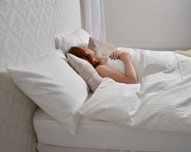Portrait of beautiful red haired woman wearing comfortable pyjamas, casualy resting in bed, getting ready for sleep,  in a glamorous white bedroom location background.