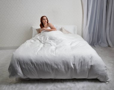 Güzel kızıl saçlı kadın portresi rahat pijama giyiyor, yatakta dinleniyor, uyumaya hazırlanıyor, göz alıcı beyaz yatak odası arka planında..