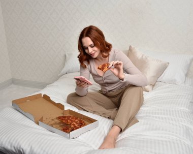 Portrait of beautiful red haired woman wearing comfortable pyjamas.Relaxing at home, eating  take out pizza,  in a  glamorous bedroom background.