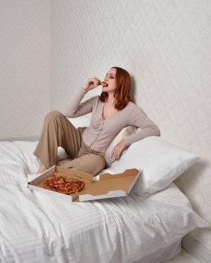 Portrait of beautiful red haired woman wearing comfortable pyjamas.Relaxing at home, eating  take out pizza,  in a  glamorous bedroom background.