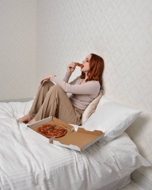 Portrait of beautiful red haired woman wearing comfortable pyjamas.Relaxing at home, eating  take out pizza,  in a  glamorous bedroom background.