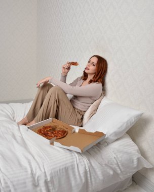 Portrait of beautiful red haired woman wearing comfortable pyjamas.Relaxing at home, eating  take out pizza,  in a  glamorous bedroom background.