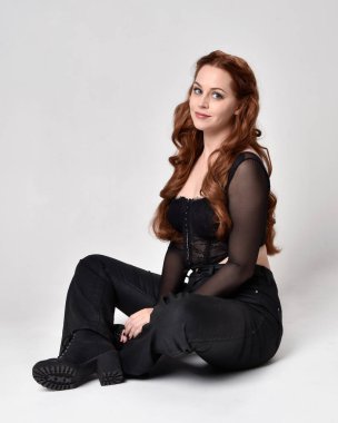 Full length portrait of beautiful woman with long red hair wearing sheer corset top and leather pants.  Sitting pose with gestural hands reaching out, kneeling on floor. Isolated on white studio background.