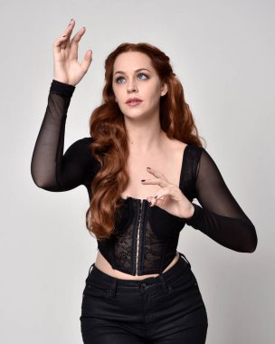 close up portrait of beautiful woman with red hair wearing sheer corset top, leather pants. Posing with gestural hands reaching out. Isolated on white studio background.