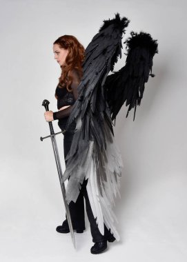 Full length portrait of beautiful woman with long red hair wearing  corset top, leather pants, large black angel feather wings. Standing pose holding sword weapon, walking forwards with gestural hands reaching out. Isolated on white studio background