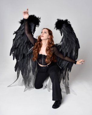 Full length portrait of beautiful woman with long red hair wearing sheer corset top, leather pants & large black angel feather wings.  Sitting pose with gestural hands reaching out, kneeling on floor. Isolated on white studio background