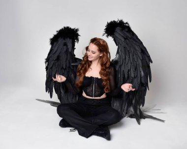 Full length portrait of beautiful woman with long red hair wearing sheer corset top, leather pants & large black angel feather wings.  Sitting pose with gestural hands reaching out, kneeling on floor. Isolated on white studio background