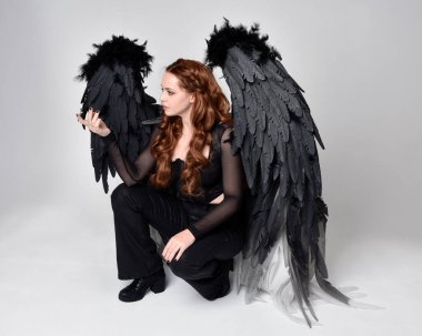 Full length portrait of beautiful woman with long red hair wearing sheer corset top, leather pants & large black angel feather wings.  Sitting pose with gestural hands reaching out, kneeling on floor. Isolated on white studio background