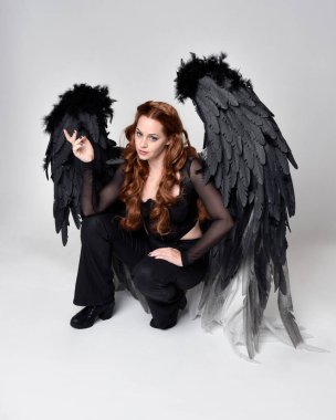 Full length portrait of beautiful woman with long red hair wearing sheer corset top, leather pants & large black angel feather wings.  Sitting pose with gestural hands reaching out, kneeling on floor. Isolated on white studio background