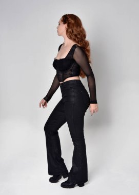 Full length portrait of beautiful woman with long red hair wearing black corset top and leather pants. Standing pose, walking forwards with gestural hands reaching out. Isolated on white studio background.