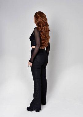 Full length portrait of beautiful woman with long red hair wearing black corset top and leather pants. Standing pose, facing backwards walking away from camera. Isolated on white studio background.