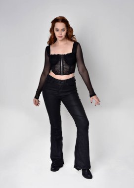 Full length portrait of beautiful woman with long red hair wearing black corset top and leather pants. Standing pose, walking forwards with gestural hands reaching out. Isolated on white studio background.