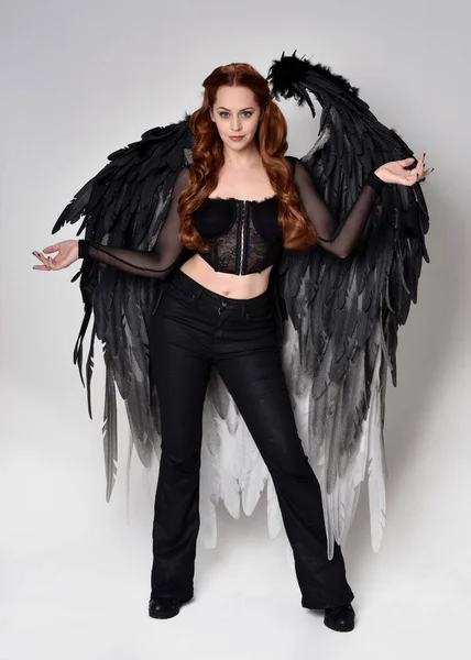 stock image Full length portrait of beautiful woman with long red hair wearing sheer corset top, leather pants, large black angel feather wings. Standing pose, walking forwards with gestural hands reaching out. Isolated on white studio background.