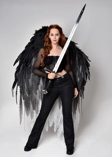 stock image Full length portrait of beautiful woman with long red hair wearing  corset top, leather pants, large black angel feather wings. Standing pose holding sword weapon, walking forwards with gestural hands reaching out. Isolated on white studio background