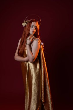 Kızıl saçlı, tanrıça ipek cüppeli ve altın kaplamalı güzel kadın modelinin fantezi portresi. El hareketleriyle poz vermek. Koyu kırmızı stüdyo arka planında izole edilmiş. 