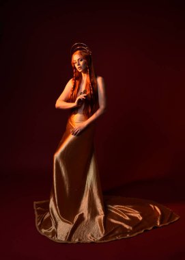 Kızıl saçlı, tanrıça ipek cüppeli ve altın kaplamalı güzel kadın modelinin tam boy fantezi portresi. Ayakta duran hareketli eller, koyu kırmızı stüdyo arka planında izole bir şekilde uzanıyor. 