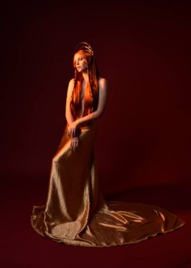 Kızıl saçlı, tanrıça ipek cüppeli ve altın kaplamalı güzel kadın modelinin tam boy fantezi portresi. Ayakta duran hareketli eller, koyu kırmızı stüdyo arka planında izole bir şekilde uzanıyor. 