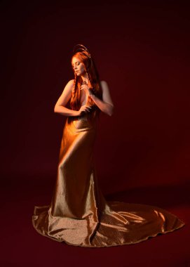 Kızıl saçlı, tanrıça ipek cüppeli ve altın kaplamalı güzel kadın modelinin tam boy fantezi portresi. Ayakta duran hareketli eller, koyu kırmızı stüdyo arka planında izole bir şekilde uzanıyor. 