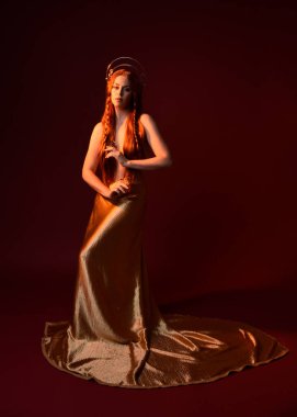 Kızıl saçlı, tanrıça ipek cüppeli ve altın kaplamalı güzel kadın modelinin tam boy fantezi portresi. Ayakta duran hareketli eller, koyu kırmızı stüdyo arka planında izole bir şekilde uzanıyor. 