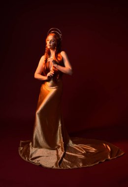 Kızıl saçlı, tanrıça ipek cüppeli ve altın kaplamalı güzel kadın modelinin tam boy fantezi portresi. Ayakta duran hareketli eller, koyu kırmızı stüdyo arka planında izole bir şekilde uzanıyor. 