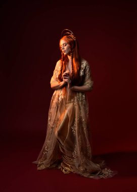 Kızıl saçlı, tanrıça ipek cüppeli ve altın kaplamalı güzel kadın modelinin tam boy fantezi portresi. Ayakta duran hareketli eller, koyu kırmızı stüdyo arka planında izole bir şekilde uzanıyor. 