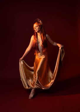 Kızıl saçlı, tanrıça ipek cüppeli ve altın kaplamalı güzel kadın modelinin tam boy fantezi portresi. Ayakta duran hareketli eller, koyu kırmızı stüdyo arka planında izole bir şekilde uzanıyor. 