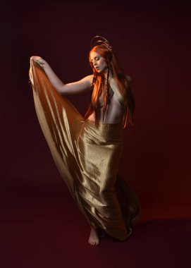 Kızıl saçlı, tanrıça ipek cüppeli ve altın kaplamalı güzel kadın modelinin tam boy fantezi portresi. Ayakta duran hareketli eller, koyu kırmızı stüdyo arka planında izole bir şekilde uzanıyor. 