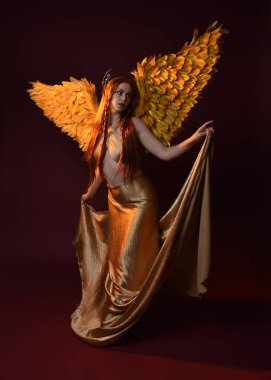 Uzun kızıl saçlı, altın ipek cüppeli, taç ve fantezi tüylü melek kanatlı güzel kadın modelinin tam boy portresi. Ayakta duran hareketli eller, koyu kırmızı stüdyo arka planında izole bir şekilde uzanıyor.