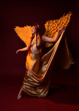 Uzun kızıl saçlı, altın ipek cüppeli, taç ve fantezi tüylü melek kanatlı güzel kadın modelinin tam boy portresi. Ayakta duran hareketli eller, koyu kırmızı stüdyo arka planında izole bir şekilde uzanıyor.