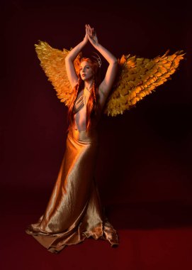 Uzun kızıl saçlı, altın ipek cüppeli, taç ve fantezi tüylü melek kanatlı güzel kadın modelinin tam boy portresi. Ayakta duran hareketli eller, koyu kırmızı stüdyo arka planında izole bir şekilde uzanıyor.