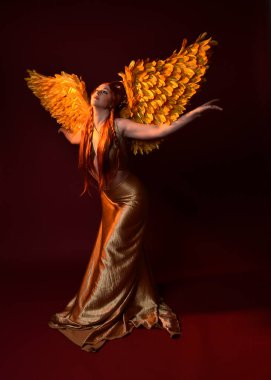 Uzun kızıl saçlı, altın ipek cüppeli, taç ve fantezi tüylü melek kanatlı güzel kadın modelinin tam boy portresi. Ayakta duran hareketli eller, koyu kırmızı stüdyo arka planında izole bir şekilde uzanıyor.
