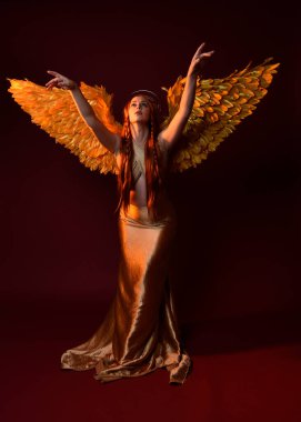 Uzun kızıl saçlı, altın ipek cüppeli, taç ve fantezi tüylü melek kanatlı güzel kadın modelinin tam boy portresi. Ayakta duran hareketli eller, koyu kırmızı stüdyo arka planında izole bir şekilde uzanıyor.