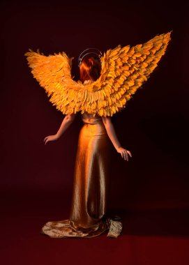 Uzun kızıl saçlı, altın ipek cüppeli, taç ve fantezi tüylü melek kanatlı güzel kadın modelinin tam boy portresi. Ayakta duran hareketli eller, koyu kırmızı stüdyo arka planında izole bir şekilde uzanıyor.