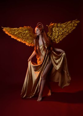 Uzun kızıl saçlı, altın ipek cüppeli, taç ve fantezi tüylü melek kanatlı güzel kadın modelinin tam boy portresi. Ayakta duran hareketli eller, koyu kırmızı stüdyo arka planında izole bir şekilde uzanıyor.