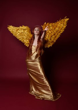 Uzun kızıl saçlı, altın ipek cüppeli, taç ve fantezi tüylü melek kanatlı güzel kadın modelinin tam boy portresi. Ayakta duran hareketli eller, koyu kırmızı stüdyo arka planında izole bir şekilde uzanıyor.