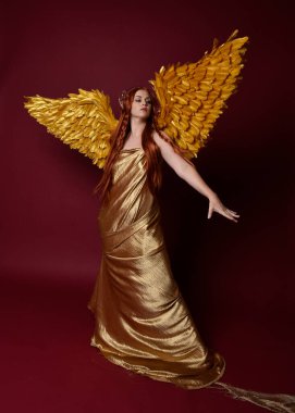 Uzun kızıl saçlı, altın ipek cüppeli, taç ve fantezi tüylü melek kanatlı güzel kadın modelinin tam boy portresi. Ayakta duran hareketli eller, koyu kırmızı stüdyo arka planında izole bir şekilde uzanıyor.