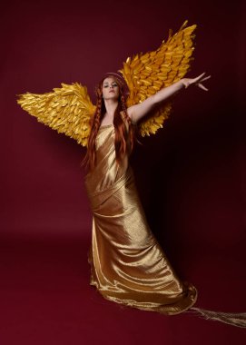 Uzun kızıl saçlı, altın ipek cüppeli, taç ve fantezi tüylü melek kanatlı güzel kadın modelinin tam boy portresi. Ayakta duran hareketli eller, koyu kırmızı stüdyo arka planında izole bir şekilde uzanıyor.