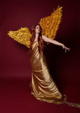 Uzun kızıl saçlı, altın ipek cüppeli, taç ve fantezi tüylü melek kanatlı güzel kadın modelinin tam boy portresi. Ayakta duran hareketli eller, koyu kırmızı stüdyo arka planında izole bir şekilde uzanıyor.