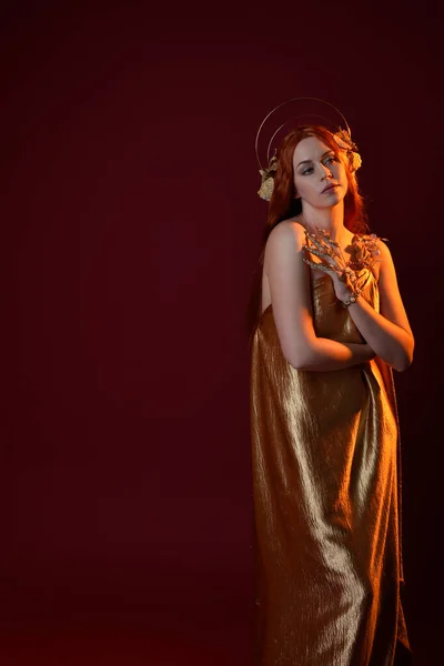 stock image Close up fantasy portrait of beautiful woman model with red hair, goddess silk robes & gold crown.  Posing with gestural hands reaching out, isolated on dark red studio background 