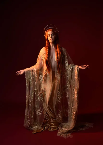 stock image Full length fantasy portrait of beautiful woman model with red hair, goddess silk robes & gold crown. Standing pose gestural hands reaching out isolated on dark red studio background 