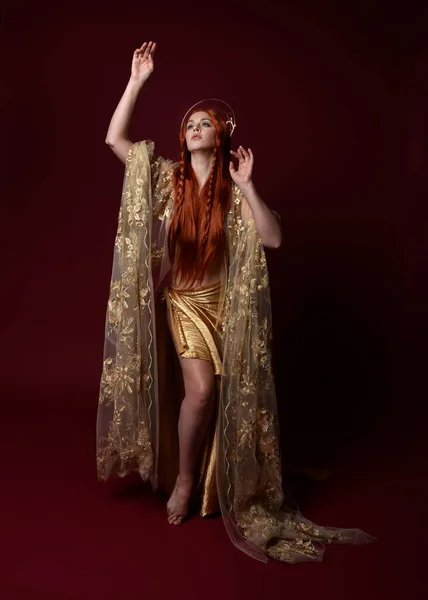 stock image Full length fantasy portrait of beautiful woman model with red hair, goddess silk robes & gold crown. Standing pose gestural hands reaching out isolated on dark red studio background 