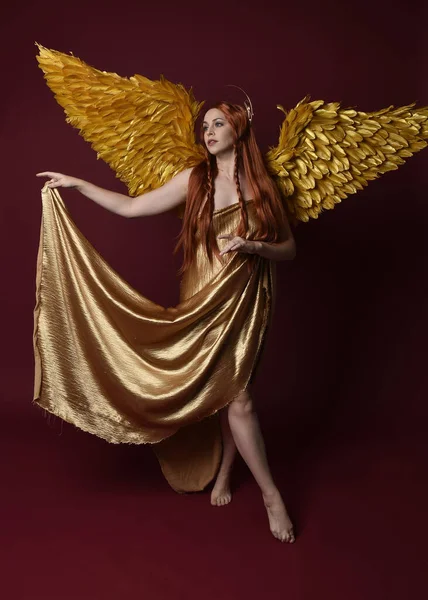 Uzun kızıl saçlı, altın ipek cüppeli, taç ve fantezi tüylü melek kanatlı güzel kadın modelinin tam boy portresi. Ayakta duran hareketli eller, koyu kırmızı stüdyo arka planında izole bir şekilde uzanıyor.