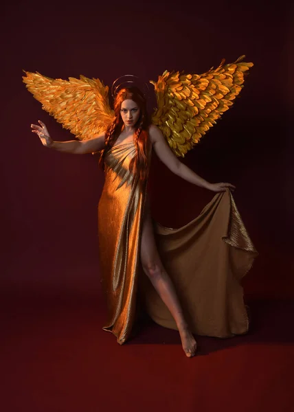 stock image Full length portrait of beautiful woman model with long red hair, gold silk robes, crown & fantasy feather angel wings. Standing pose gestural hands reaching out isolated on dark red studio background