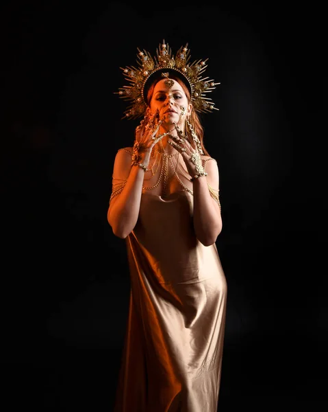 Close up fantasy portrait of beautiful woman model with red hair, goddess silk robes & ornate gold crown.  Posing with gestural hands reaching out, isolated on dark  studio background