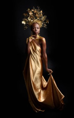  Afro, tanrıça ipek cübbesi ve süslü çiçek tacıyla güzel Afrikalı kadın modelinin fantezi portresi. Altın çiçekleri tutan hareketli pozlar. Karanlık stüdyo arka planında izole 