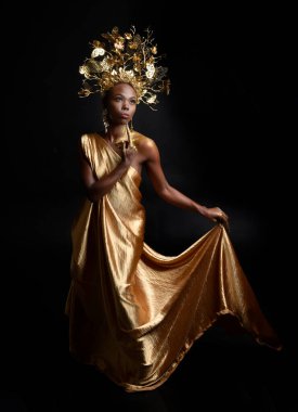  Afro, tanrıça ipek cübbesi ve süslü çiçek tacıyla güzel Afrikalı kadın modelinin fantezi portresi. Altın çiçekleri tutan hareketli pozlar. Karanlık stüdyo arka planında izole 