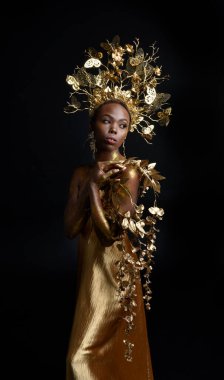  Afro, tanrıça ipek cübbesi ve süslü çiçek tacıyla güzel Afrikalı kadın modelinin fantezi portresi. Altın çiçekleri tutan hareketli pozlar. Karanlık stüdyo arka planında izole 