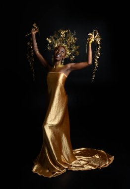  Afro, tanrıça ipek cübbesi ve süslü çiçek tacıyla güzel Afrikalı kadın modelinin fantezi portresi. Altın çiçekleri tutan hareketli pozlar. Karanlık stüdyo arka planında izole 