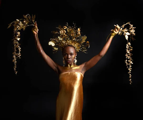 アフロ 女神シルクのローブと華やかな花冠と美しいアフリカの女性モデルの幻想的な肖像画 ジェスチャー黄金の花を保持ポーズ 暗いスタジオの背景に隔離され — ストック写真