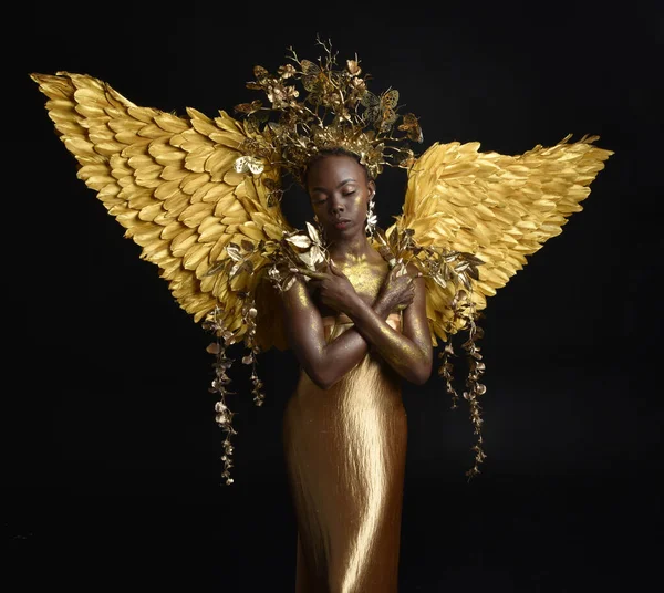 fantasy portrait of beautiful african woman model with afro, goddess silk robes, ornate crown & gold angel wings.  gestural Posing holding golden flowers., isolated on dark  studio background
